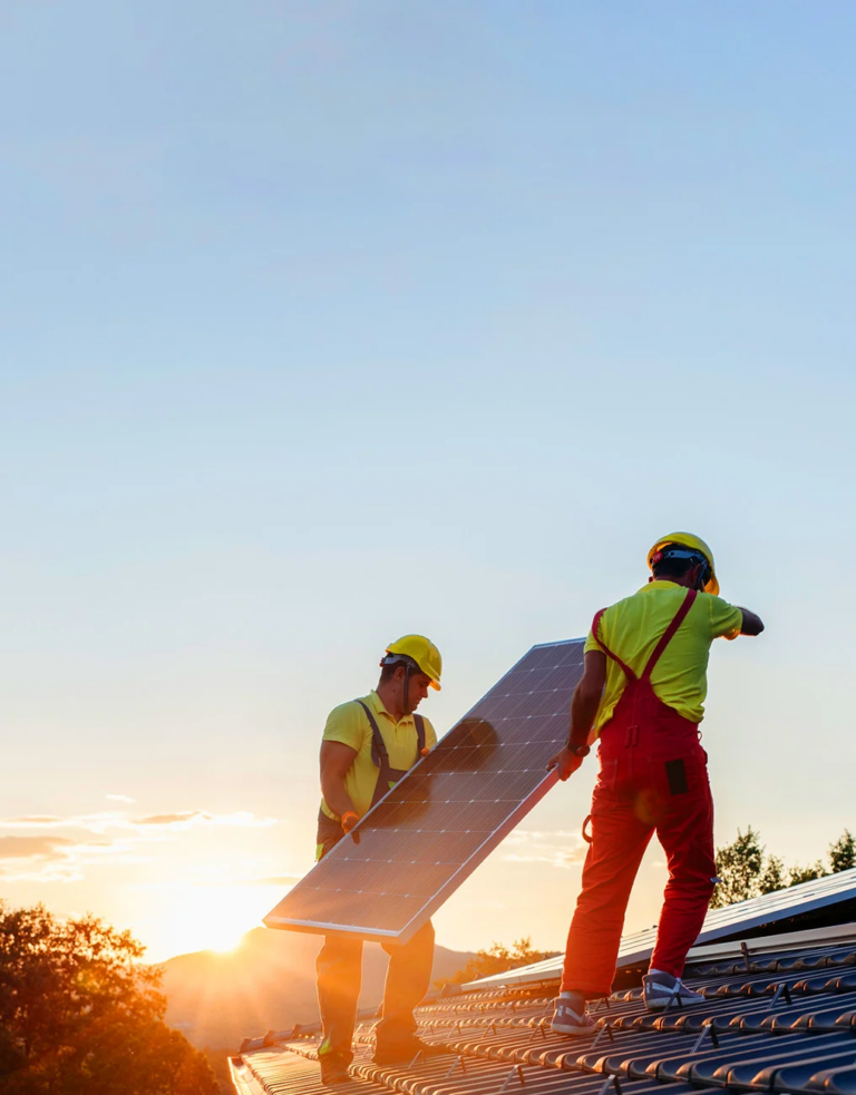 To arbeidere som bærer et solcellepanel som skal installeres på et svart tak. Solen skinner på mennene mens den går ned bak noen fjell i bakgrunnen. Foto